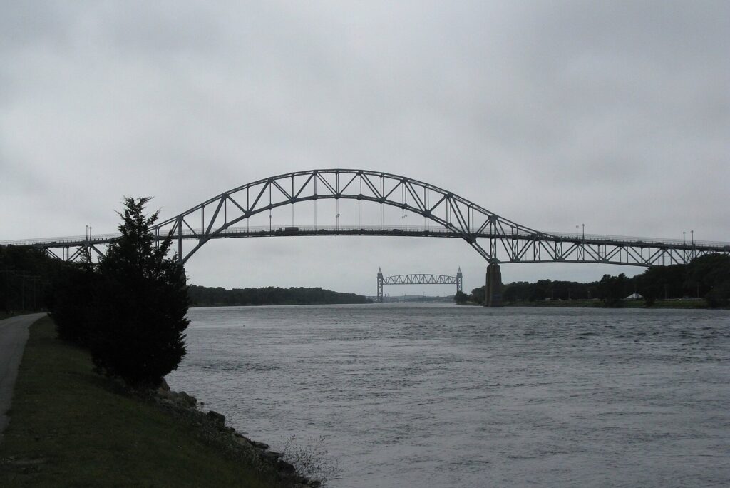 The steel arch of the Bourne Bridge over the Cape Cod Canal under a gray sky, with the railroad bridge in the distance