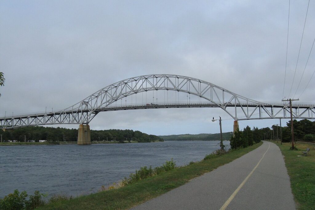 A paved path running beside the Cape Cod Canal toward the steel arch of the Bourne Bridge under a cloudy sky