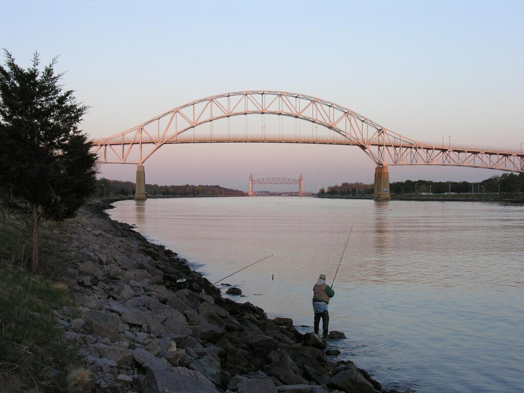 An angler casting from the rocky bank of the Cape Cod Canal at sunset, with the Bourne Bridge arching over the water
