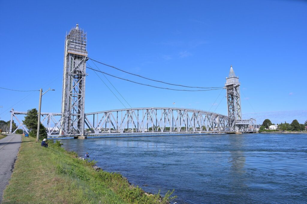 Anglers on the grassy bank of the Cape Cod Canal beside the tall lift towers of the railroad bridge on a clear day