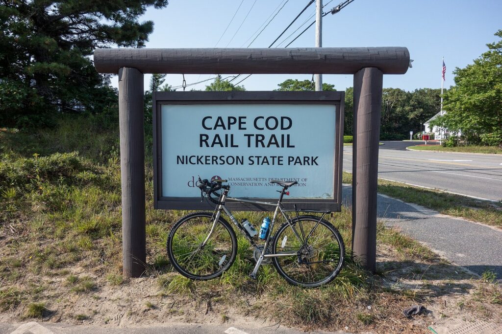 A wooden-framed Cape Cod Rail Trail sign reading Nickerson State Park, with a bicycle leaning against it beside the path
