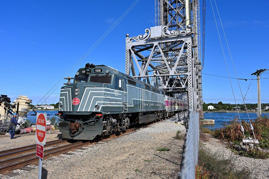 The CapeFLYER train, led by a gray and green locomotive, crossing the Cape Cod Canal Railroad Bridge on a sunny day