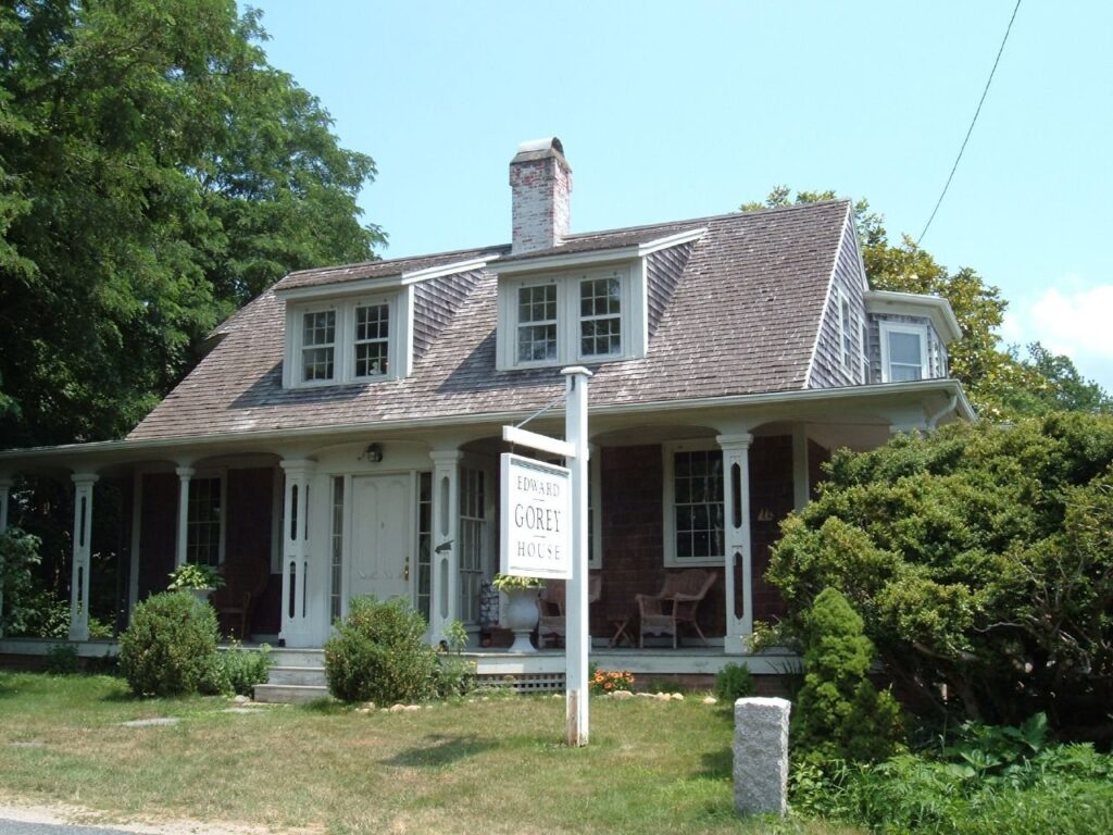 The shingled Edward Gorey House with its porch and signpost among green shrubs in Yarmouth Port