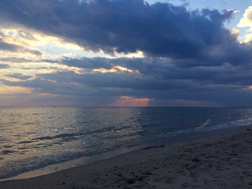 Sunset breaking through heavy clouds over calm water and a wide sandy beach at Herring Cove in Provincetown
