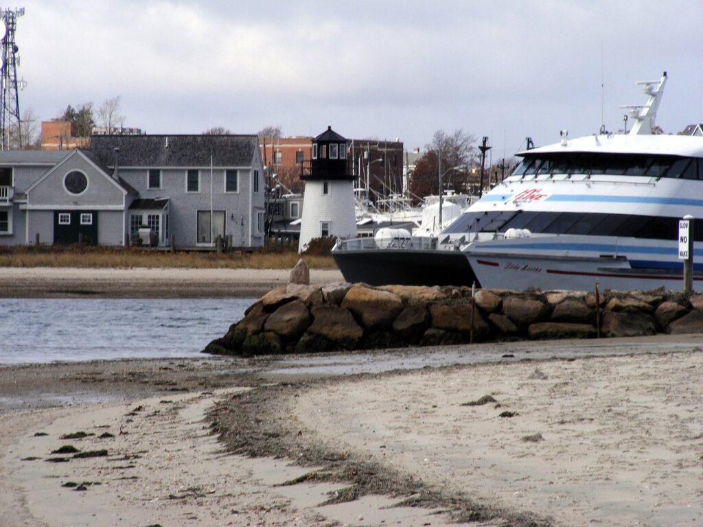 A white high-speed ferry docked in Hyannis Inner Harbor beside a small white lighthouse and a sandy beach