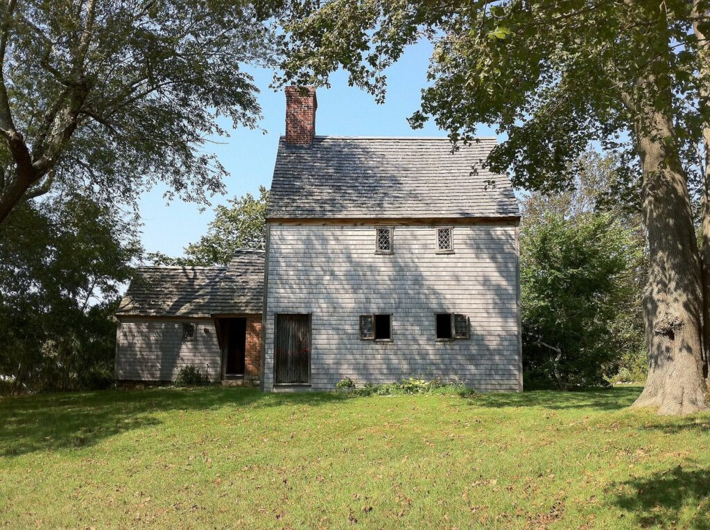 The gray-shingled Hoxie House, a saltbox with a steep roof and a central chimney, on a lawn under large trees in Sandwich