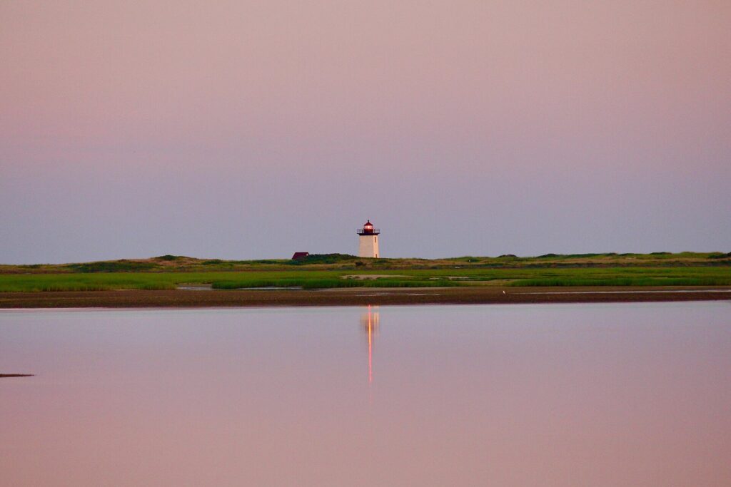 Long Point Lighthouse on a low strip of land reflected in still water under a pink sunset sky in Provincetown Harbor
