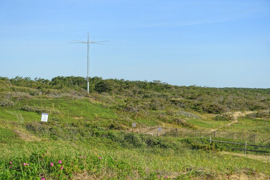 Low green heathland and dune scrub at the Marconi Station Site in South Wellfleet, with a tall antenna mast under a blue sky