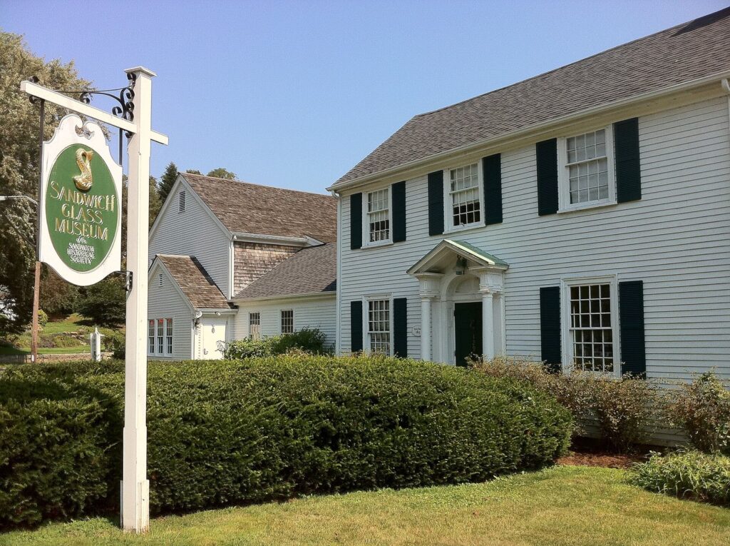 The white clapboard Sandwich Glass Museum with dark shutters and its hanging sign on a sunny day