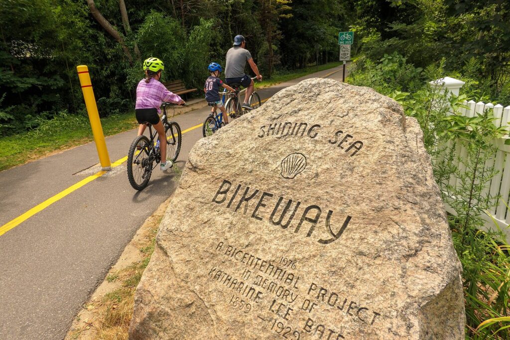 A carved boulder marking the Shining Sea Bikeway beside the paved path as a family rides past on bicycles