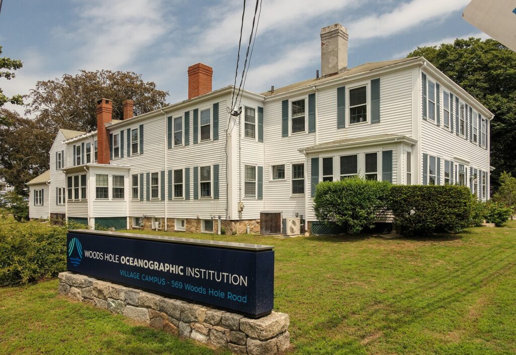 A white clapboard building behind the Woods Hole Oceanographic Institution sign on Woods Hole Road